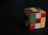 A partially solved Rubik's Cube with various colors including red, green, yellow, and blue on a dark surface. The cube's orientation emphasizes its three-dimensional shape, with a slight shadow cast.