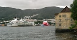 A waterfront scene featuring two large ships docked in a harbor with a backdrop of lush green hills and a town with clustered white and red-roofed houses. A prominent wooden building with peeling yellow paint stands on the right, partially overhanging the water.
