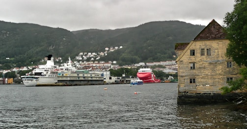 A waterfront scene featuring two large ships docked in a harbor with a backdrop of lush green hills and a town with clustered white and red-roofed houses. A prominent wooden building with peeling yellow paint stands on the right, partially overhanging the water.