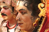 Two individuals are dressed in traditional Indian theatrical costumes with elaborate makeup and headpieces featuring intricate designs and vibrant colors. The focus is on their facial expressions, characterized by bold eyeliner and facial adornments. The attire includes jewel-toned ornaments and embroidered details.