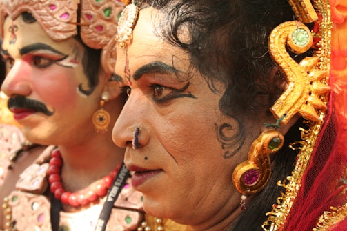 Two individuals are dressed in traditional Indian theatrical costumes with elaborate makeup and headpieces featuring intricate designs and vibrant colors. The focus is on their facial expressions, characterized by bold eyeliner and facial adornments. The attire includes jewel-toned ornaments and embroidered details.