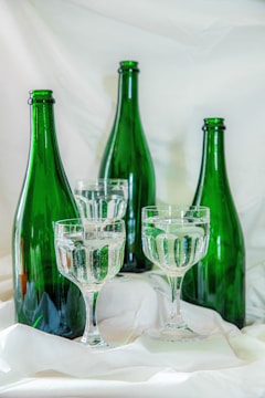 Four green glass bottles are arranged alongside two clear stemmed glasses filled with a transparent liquid. They are set against a soft, white fabric background that creates a gentle and elegant scene.