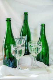 Four green glass bottles are arranged alongside two clear stemmed glasses filled with a transparent liquid. They are set against a soft, white fabric background that creates a gentle and elegant scene.