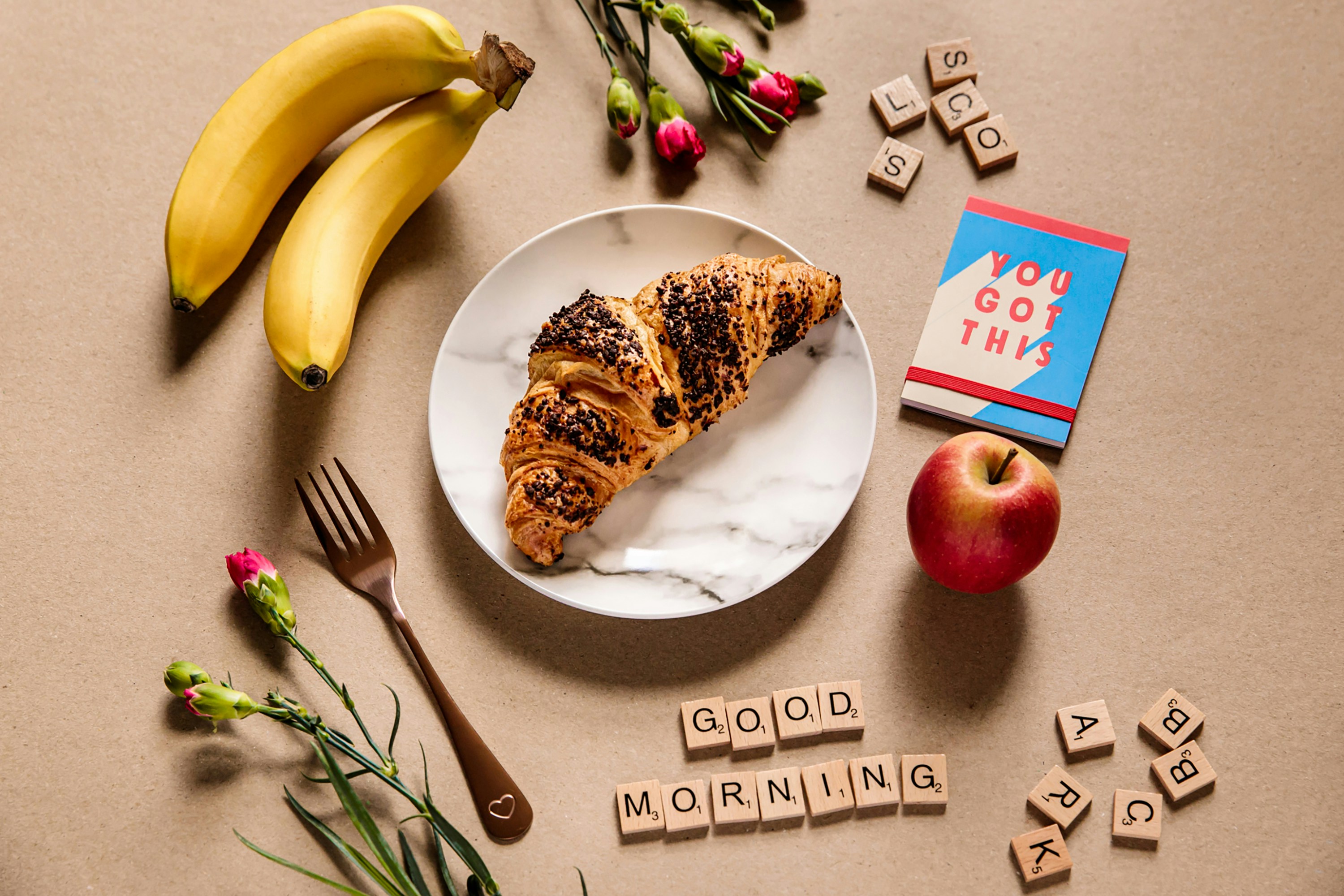 A croissant on a marble plate with a gold fork, apple, bananas, roses, You Got This notebook and Good Morning scrabble tiles