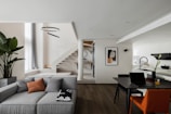 A modern and stylish interior space featuring a spacious living area with a grey sectional sofa, enhanced by a few decorative cushions and a blanket. There's a potted plant adding a touch of greenery next to the couch. The dining area includes a contemporary black table with an orange chair and a laptop, suggesting an office-like setting. A staircase with wooden steps leads to an upper level. Above, a circular light fixture adds an elegant touch. The kitchen area is bright and minimalistic, with white cabinetry and a sleek design. A piece of abstract art decorates the wall next to the stairs.