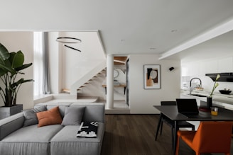 A modern and stylish interior space featuring a spacious living area with a grey sectional sofa, enhanced by a few decorative cushions and a blanket. There's a potted plant adding a touch of greenery next to the couch. The dining area includes a contemporary black table with an orange chair and a laptop, suggesting an office-like setting. A staircase with wooden steps leads to an upper level. Above, a circular light fixture adds an elegant touch. The kitchen area is bright and minimalistic, with white cabinetry and a sleek design. A piece of abstract art decorates the wall next to the stairs.