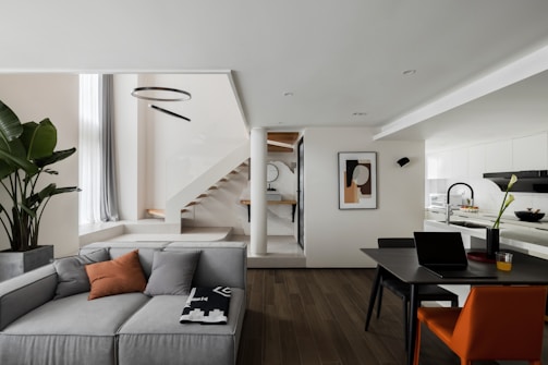 A modern and stylish interior space featuring a spacious living area with a grey sectional sofa, enhanced by a few decorative cushions and a blanket. There's a potted plant adding a touch of greenery next to the couch. The dining area includes a contemporary black table with an orange chair and a laptop, suggesting an office-like setting. A staircase with wooden steps leads to an upper level. Above, a circular light fixture adds an elegant touch. The kitchen area is bright and minimalistic, with white cabinetry and a sleek design. A piece of abstract art decorates the wall next to the stairs.