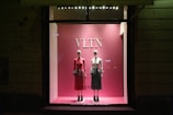 A pink-themed store display window features two mannequins dressed in stylish outfits. The mannequin on the left is wearing a long-sleeved pink dress with a black handbag, while the mannequin on the right is dressed in a white top and a green skirt with a similar bag. The display is minimalist with a focus on elegance and fashion.
