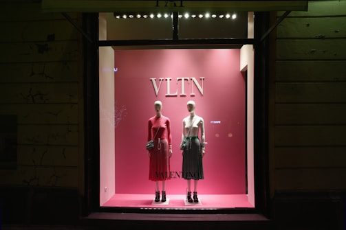 A pink-themed store display window features two mannequins dressed in stylish outfits. The mannequin on the left is wearing a long-sleeved pink dress with a black handbag, while the mannequin on the right is dressed in a white top and a green skirt with a similar bag. The display is minimalist with a focus on elegance and fashion.