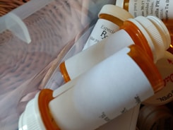 A collection of orange prescription pill bottles with white caps, some lying on their sides in a clear plastic container. Labels with printed text are visible on the bottles.