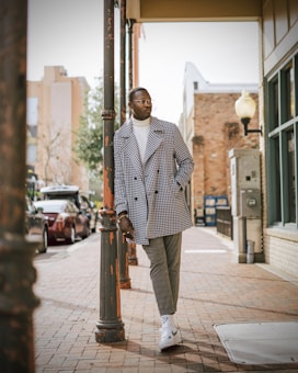 A stylishly dressed person is leaning against a street lamp on a paved sidewalk in an urban setting. They are wearing a patterned coat, white turtleneck, gray pants, and white sneakers. The background shows buildings, trees, and parked cars.