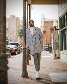 A stylishly dressed person is leaning against a street lamp on a paved sidewalk in an urban setting. They are wearing a patterned coat, white turtleneck, gray pants, and white sneakers. The background shows buildings, trees, and parked cars.