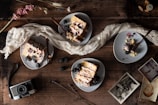 Four plates of cake garnished with blackberries and cream are arranged on a rustic wooden table, accompanied by an old-fashioned camera, two vintage photographs, and dried flowers. A white cloth is artistically draped around the plates, adding an elegant touch to the setup.