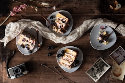 Four plates of cake garnished with blackberries and cream are arranged on a rustic wooden table, accompanied by an old-fashioned camera, two vintage photographs, and dried flowers. A white cloth is artistically draped around the plates, adding an elegant touch to the setup.
