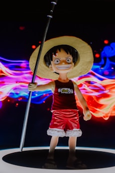 A figurine of a character wearing a straw hat, red tank top, and red shorts with white trim, wielding a long pole. The figure stands against a vibrant, swirling background of colorful lights in red, blue, and purple hues, creating a dynamic and lively scene.