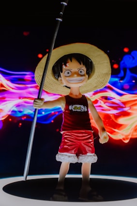 A figurine of a character wearing a straw hat, red tank top, and red shorts with white trim, wielding a long pole. The figure stands against a vibrant, swirling background of colorful lights in red, blue, and purple hues, creating a dynamic and lively scene.