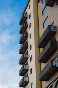 A modern apartment building with multiple balconies aligned vertically on its side. The building features a mix of beige and brown tones with metal railings on the balconies. The sky is clear with a soft blue color and scattered clouds, creating a peaceful atmosphere.