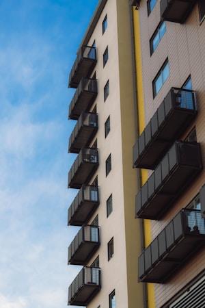 A modern apartment building with multiple balconies aligned vertically on its side. The building features a mix of beige and brown tones with metal railings on the balconies. The sky is clear with a soft blue color and scattered clouds, creating a peaceful atmosphere.