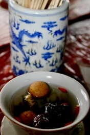 A porcelain container adorned with blue dragon patterns is filled with wooden chopsticks. In front of it, a bowl with a clear broth contains various ingredients, including round brown and dark objects, possibly jujubes and lotus seeds.