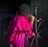 A person with dreadlocks is singing into a microphone on stage. They are wearing a vibrant pink, ruffled cloak over a dark outfit.