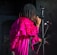 A person with dreadlocks is singing into a microphone on stage. They are wearing a vibrant pink, ruffled cloak over a dark outfit.