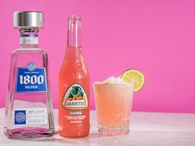 A bottle of 1800 Silver Tequila stands next to a Jarritos guava soda bottle, both placed on a marble surface. Beside them, a glass filled with a pink beverage, topped with coconut shavings and garnished with a lime slice. The background is a vibrant pink.