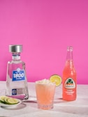 A bottle of 1800 Silver tequila and a Jarritos guava soda are positioned on a marble surface against a vibrant pink backdrop. Between them, a cocktail in a textured glass features a garnish of coconut flakes and a slice of lime. A plate with lime slices rests next to the tequila bottle.