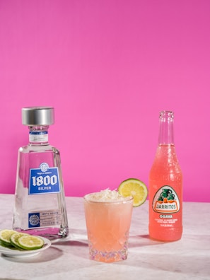 A bottle of 1800 Silver tequila and a Jarritos guava soda are positioned on a marble surface against a vibrant pink backdrop. Between them, a cocktail in a textured glass features a garnish of coconut flakes and a slice of lime. A plate with lime slices rests next to the tequila bottle.