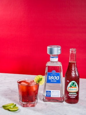 A glass filled with a red beverage, ice, and a slice of lime is placed on a marble surface. Beside it, there is a bottle of 1800 Silver Tequila and a bottle of Jarritos Jamaica soda. A few lime slices are on the table in the foreground. The background is a bold red color.