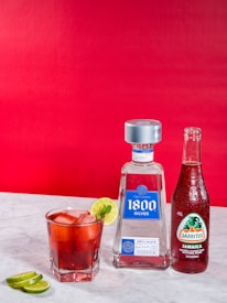 A glass filled with a red beverage, ice, and a slice of lime is placed on a marble surface. Beside it, there is a bottle of 1800 Silver Tequila and a bottle of Jarritos Jamaica soda. A few lime slices are on the table in the foreground. The background is a bold red color.
