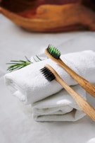 Two bamboo toothbrushes with black bristles are placed on rolled-up white towels. A sprig of rosemary adds a touch of greenery to the scene, and a wooden bowl is subtly visible in the blurred background.