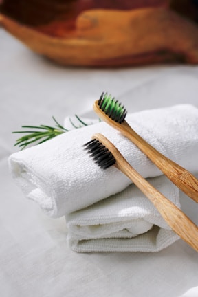 Two bamboo toothbrushes with black bristles are placed on rolled-up white towels. A sprig of rosemary adds a touch of greenery to the scene, and a wooden bowl is subtly visible in the blurred background.