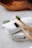 Two bamboo toothbrushes with black bristles are placed on rolled-up white towels. A sprig of rosemary adds a touch of greenery to the scene, and a wooden bowl is subtly visible in the blurred background.