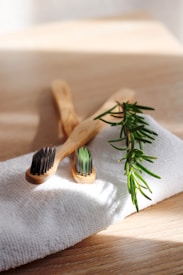 Two bamboo toothbrushes lie on a textured white cloth beside a sprig of green rosemary on a light wooden surface. Gentle sunlight streams across the scene, casting soft shadows.