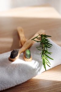 Two bamboo toothbrushes lie on a textured white cloth beside a sprig of green rosemary on a light wooden surface. Gentle sunlight streams across the scene, casting soft shadows.