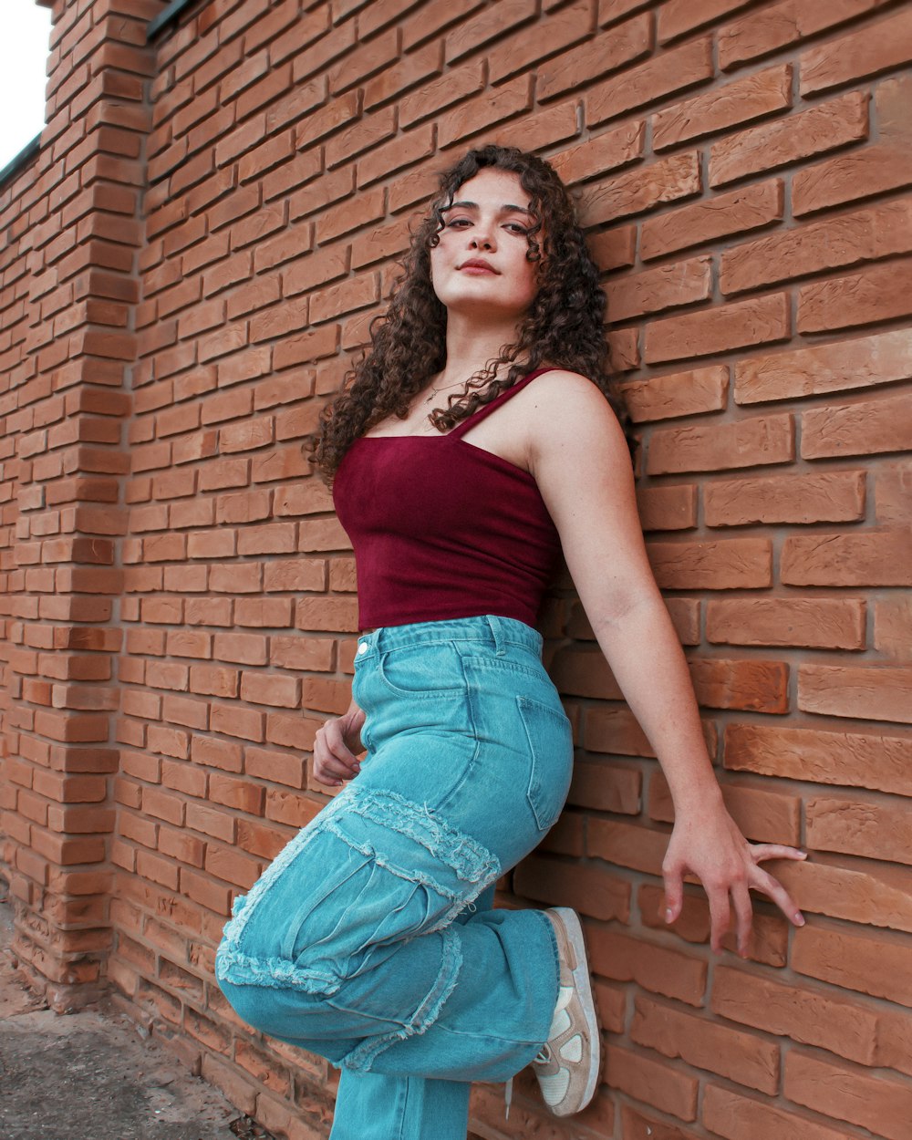 Femme en débardeur rouge et short en jean bleu assise sur un mur de briques brunes