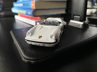 A small die-cast model car with a white exterior is placed on a black surface. In the background, several colorful books are stacked flat, providing depth and context to the setting.