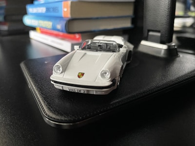 A small die-cast model car with a white exterior is placed on a black surface. In the background, several colorful books are stacked flat, providing depth and context to the setting.