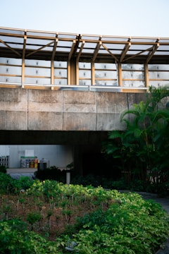 A concrete structure with a modern architectural design featuring large windows and metal beams. There is a lush garden with various green plants in the foreground. On the left side, there are visible recycling bins. The overall scene is well-lit with natural light.