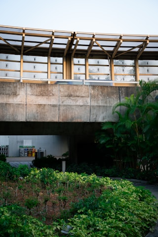 A concrete structure with a modern architectural design featuring large windows and metal beams. There is a lush garden with various green plants in the foreground. On the left side, there are visible recycling bins. The overall scene is well-lit with natural light.