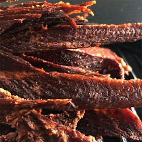 A close-up view of several strips of jerky, appearing savory and well-seasoned. The surface texture is rugged and slightly oily, consistent with dried and spiced meat. The jerky pieces are arranged closely together.