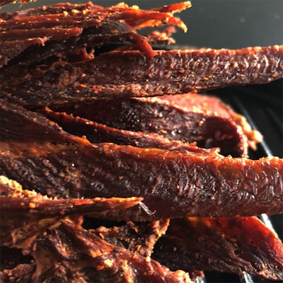 A close-up view of several strips of jerky, appearing savory and well-seasoned. The surface texture is rugged and slightly oily, consistent with dried and spiced meat. The jerky pieces are arranged closely together.