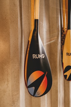 A close-up of a paddle with a shiny, carbon fiber-like blade featuring vibrant graphic designs and the word 'RUMO'. The blade design includes a circle with gradient colors of orange and blue, and a stylized arrow. The paddle's handle is wooden, and it is leaning against a textured wall.