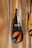 A close-up of a paddle with a shiny, carbon fiber-like blade featuring vibrant graphic designs and the word 'RUMO'. The blade design includes a circle with gradient colors of orange and blue, and a stylized arrow. The paddle's handle is wooden, and it is leaning against a textured wall.