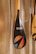 A close-up of a paddle with a shiny, carbon fiber-like blade featuring vibrant graphic designs and the word 'RUMO'. The blade design includes a circle with gradient colors of orange and blue, and a stylized arrow. The paddle's handle is wooden, and it is leaning against a textured wall.