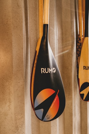 A close-up of a paddle with a shiny, carbon fiber-like blade featuring vibrant graphic designs and the word 'RUMO'. The blade design includes a circle with gradient colors of orange and blue, and a stylized arrow. The paddle's handle is wooden, and it is leaning against a textured wall.