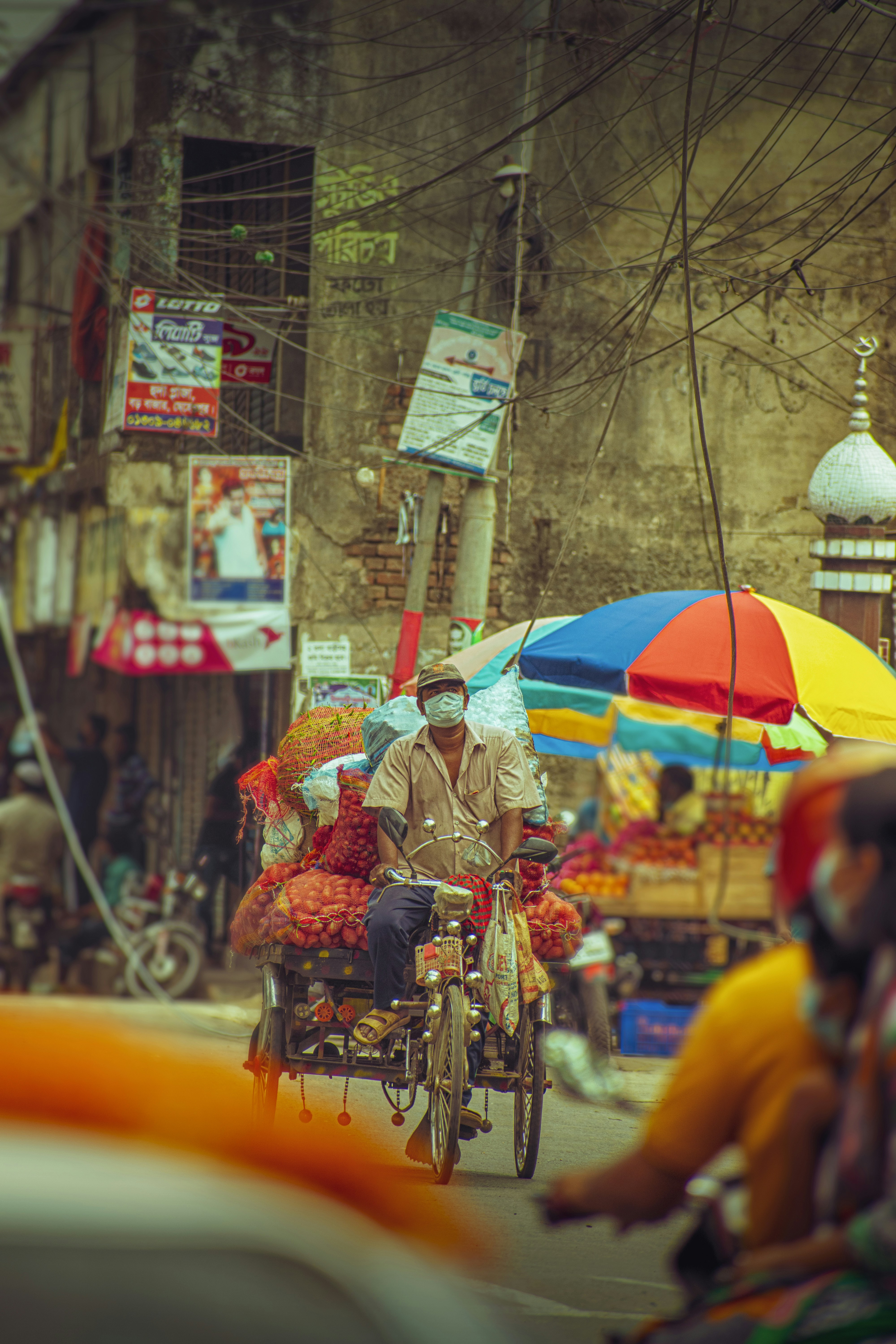 A busy market street with colorful umbrellas and a cycle rickshaw loaded with goods.