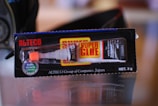 A tube of super glue in its packaging. The package is black with red and yellow accents and displays the brand name 'Alteco' prominently. The glue tube is visible through a clear section of the packaging, with a white and orange dispensing nozzle.