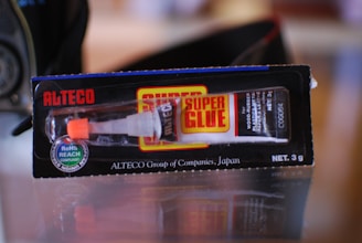A tube of super glue in its packaging. The package is black with red and yellow accents and displays the brand name 'Alteco' prominently. The glue tube is visible through a clear section of the packaging, with a white and orange dispensing nozzle.