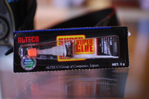 A tube of super glue in its packaging. The package is black with red and yellow accents and displays the brand name 'Alteco' prominently. The glue tube is visible through a clear section of the packaging, with a white and orange dispensing nozzle.